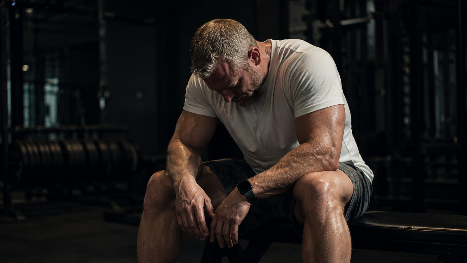 A fit person sitting alone on a gym bench with head bowed and hands on knees, expression of genuine exhaustion rather than post-workout satisfaction, dim gym lighting, representing the moment of recognizing overtraining before it forces a complete stop