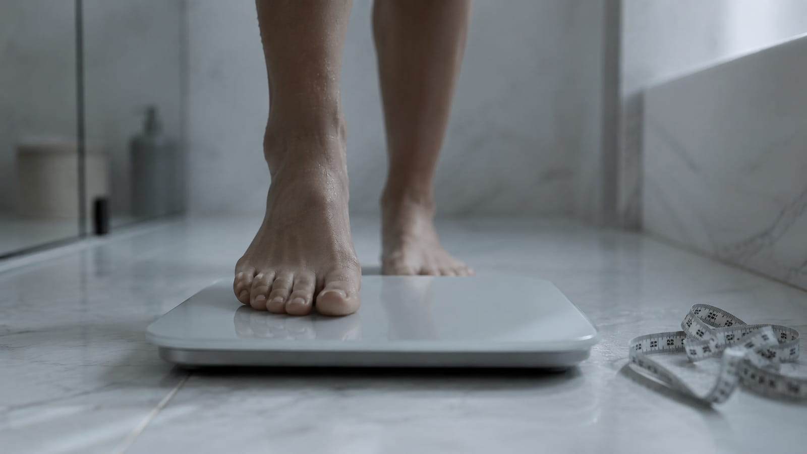 A person stepping onto a bathroom scale with a measuring tape nearby, representing the incomplete picture that scale weight alone tells about body composition and health during GLP-1 medication use