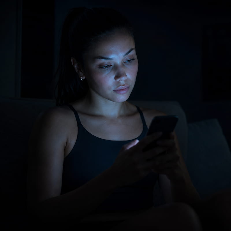 An athletic woman sitting alone in a dark room illuminated only by the glow of a smartphone screen, passive and unfocused expression, representing a mind being directed by outside forces rather than intentional thought